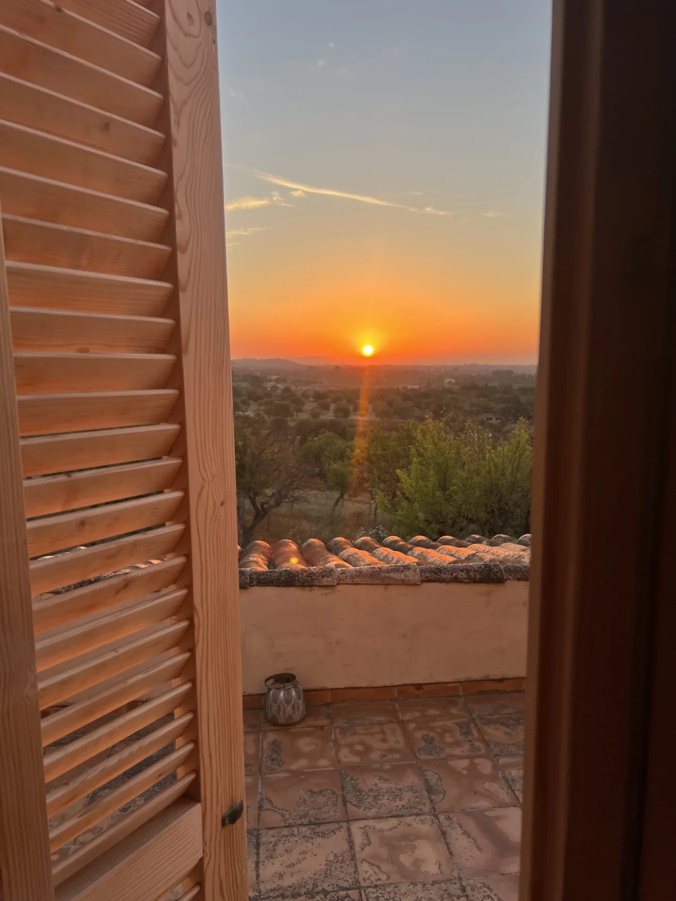 Den oransje solen går ned over Mallorca, sett gjennom husets åpne skodder